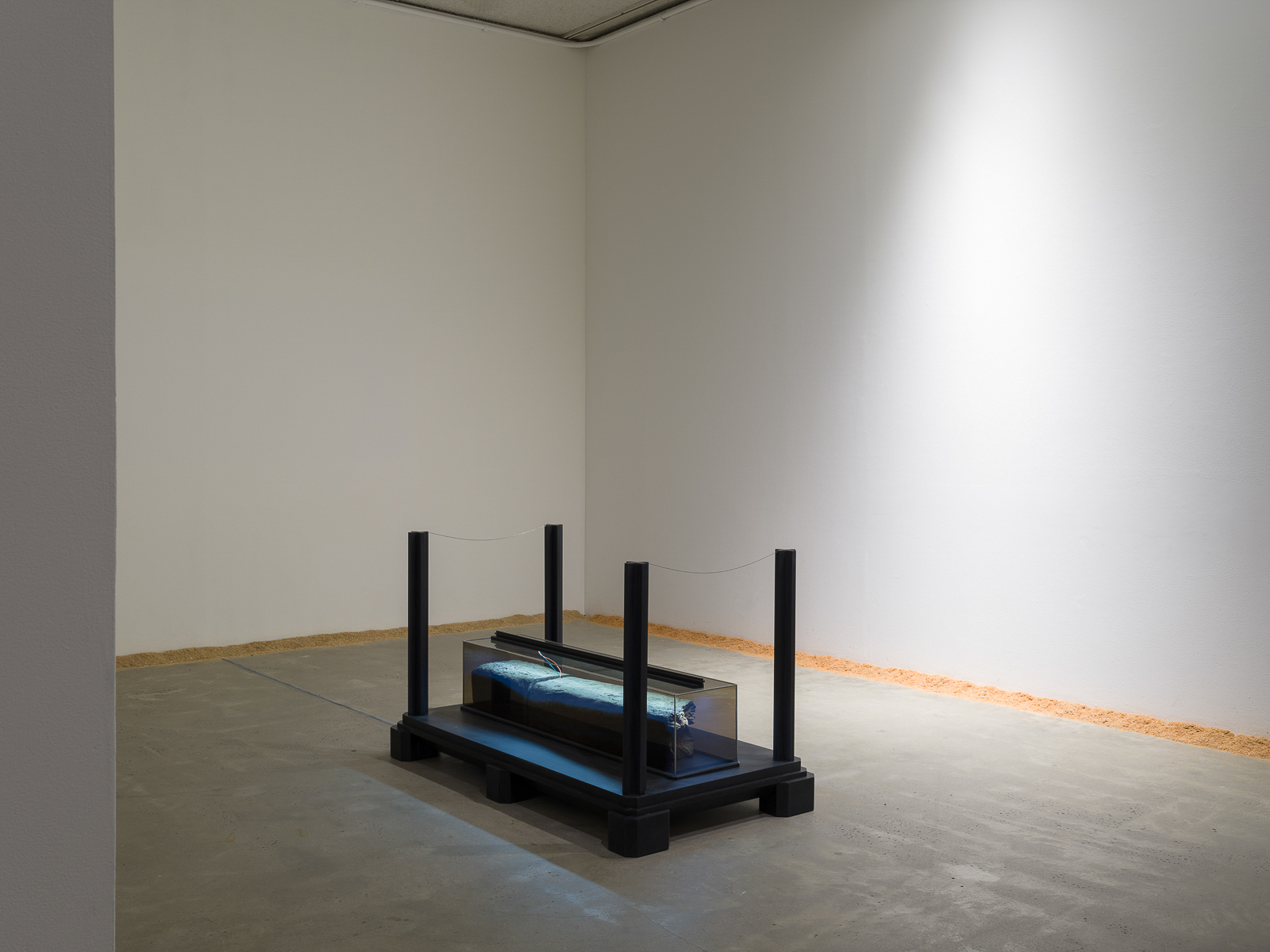 This is a color photograph of two artworks by Lukas Geronimas. In the foreground is the sculpture entitled Mayfair Column Remainder, which is depicted at a slight angle. It has been placed on the floor, centered in the room. It consists of a rectangular black platform with four black posts in each corner. Centered on the platform is a rectangular column on its side, made of transparent plexiglass containing a railroad tie, which is a rectangular piece of wood. It is illuminated with an LED light. In the background to the right of the sculpture and along the perimeter of the floor and walls of this installation is another artwork titled Puzzledust, which is sawdust that was produced, collected and stored by the artist over the past two years.