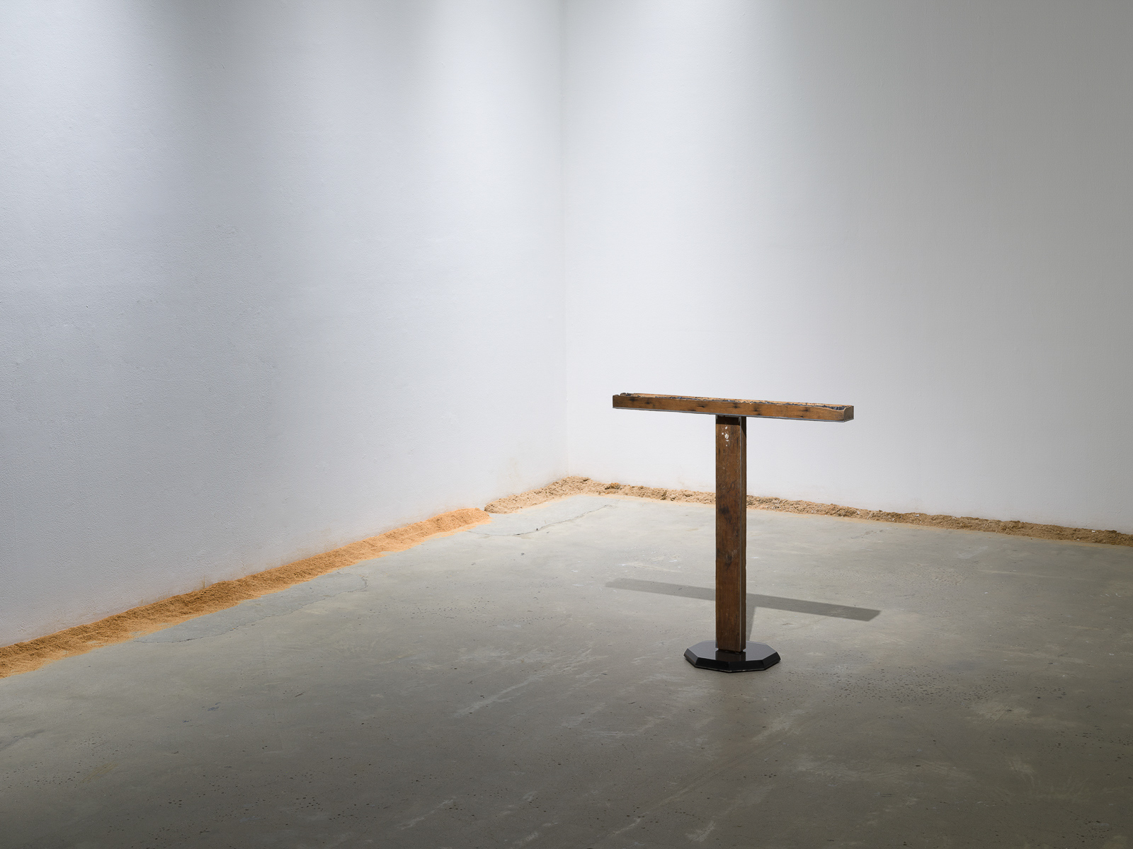 This is a color photograph of two artworks by Lukas Geronimas. In the foreground to the right and featured at a slight angled view is a sculpture entitled Custom T, composed of two rectangular pieces of wood with one mounted perpendicular on top of the other to form the shape of a T. The wooden T is mounted to a black octagonal plexiglass base. In the background and along the perimeter of the floor and walls is another artwork titled Puzzledust, which is sawdust that was produced, collected and stored by the artist over the past two years.