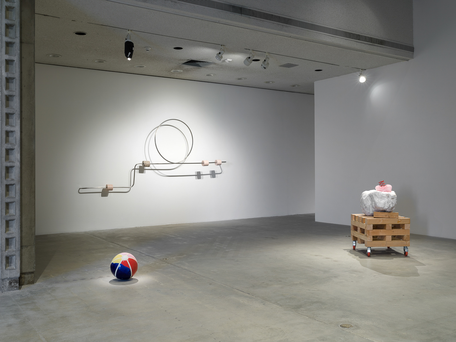 This is a color photograph of three sculptures by Nevine Mahmoud. Pictured left to right are: a sculpture titled Beach Ball IV, which is a multi-colored spherical ceramic work placed on the floor and resembles an inflatable beach ball; a wall-mounted sculpture titled Abacus arm 1, which features a linear steel bar with a circle and four stone components. Each of these components are wrapped around the steel bar and are different stones, such as onyx, Portuguese marble and Aphrodite marble; and a sculpture titled Three Components, which includes a large white boulder with a small, pink, bulbous glass object that has a red coil placed on top of it. These particular elements sit on top of a large stack of wood pieces on wheels.