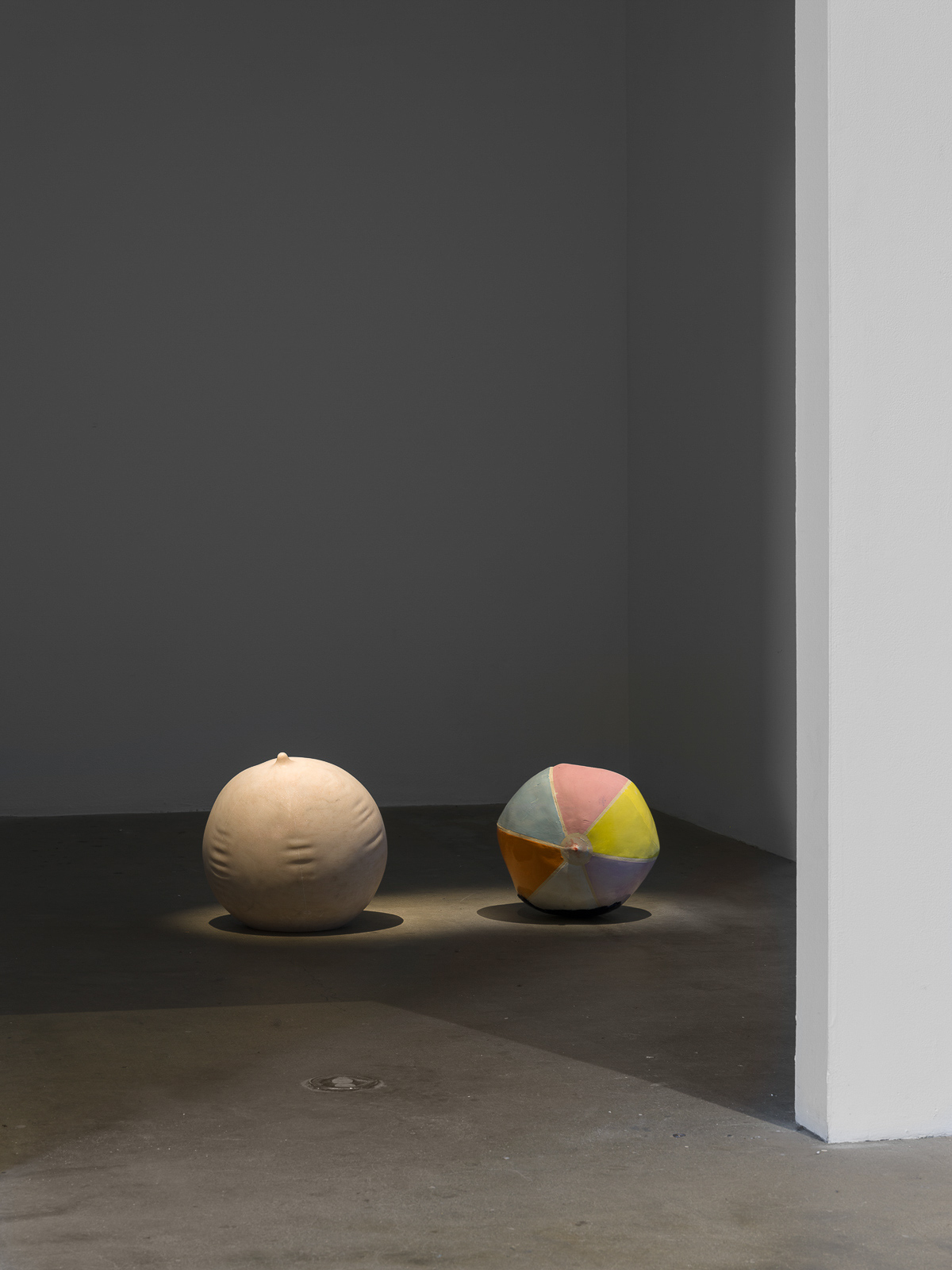 This is a color photograph of two sculptures by Nevine Mahmoud. The sculpture on the left is Ball Stripped Bare, which is spherical, made of Portuguese pink marble, and resembles an inflated beach ball. To the right of this work is Deflating Beach Ball II, which is slightly smaller in comparison to the other work and is made of ceramic with a multicolored in colors of pink, light blue, yellow and orange glaze mimicking the look of a slightly deflated beach ball. Both sculptures are placed directly on the floor and side-by-side.