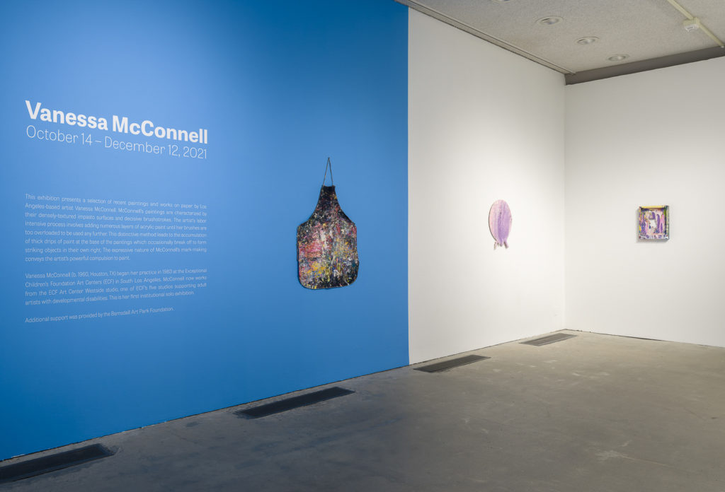 This is a color photograph of an exhibition room at the Los Angeles Municipal Art Gallery. The far left wall is painted blue and depicts an introductory wall text for Vanessa McConnell’s exhibition in white text over blue paint. To the left of this wall text is an untitled artwork, which is a dark colored apron hung flat on the wall covered in smears and splatters of bright colored paint. This artwork is installed on the blue portion of the wall. To the left of this is another artwork hung on the white portion of the wall. This artwork is an abstract painting by Vanessa McConnell, and is a circular, painted canvas with thick layers of pink and purple-colored paints. On the left adjoining wall is another artwork by the same artist, which is an abstract painting on the backside of a rectangular canvas with thick layers of paint in various colors, such as purple, pink, and green. This artwork has a hole in the middle of it.