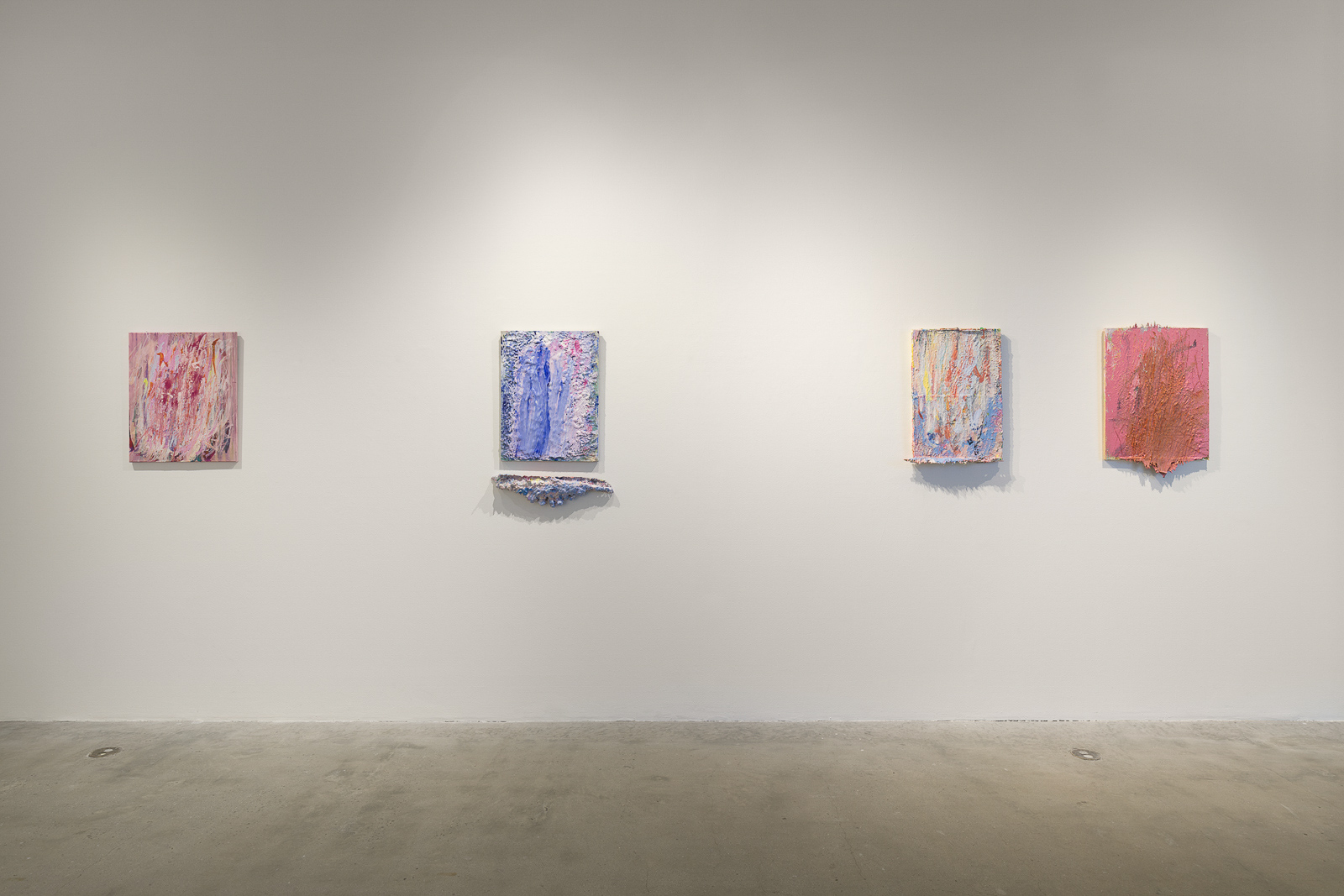 This is a color photograph of a white gallery wall with four hung artworks by Vanessa McConnell. Each artwork is an abstract painting of roughly equal size. On the far left is a painting utilizing mostly white, pink, and red paint, applied thickly to the canvas in mostly vertical strokes. In the left center is a painting consisting of mostly blue shades of paint with some pink and red as well, applied thickly to the canvas. A separate irregularly shaped piece in a similar color scheme is hung directly underneath. In the right center is a painting consisting of various colors, including yellow, blue, and red in mostly vertical strokes. On the far right is a painting featuring thick, textured layers of pink and orange paint, which extends and stretches outward from the bottom of the canvas.