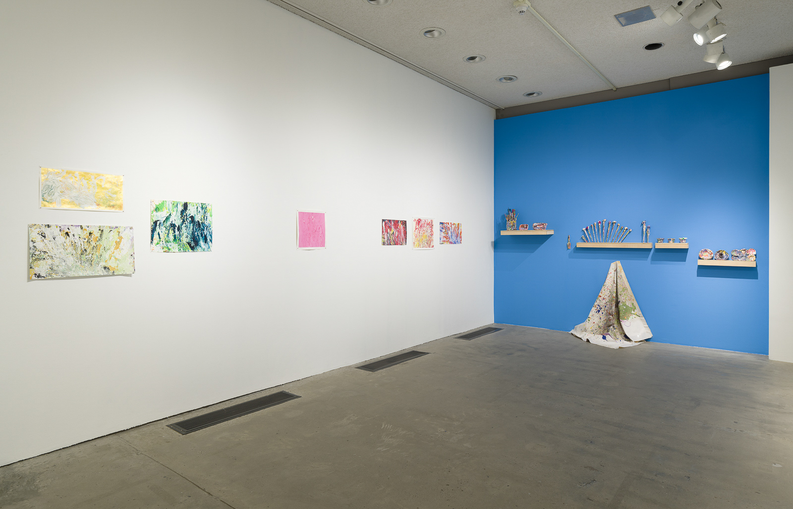 This is a color photograph of two walls in an exhibition of Vanessa McConnell's artworks at the Los Angeles Municipal Art Gallery. On the right white wall are seven brightly colored abstract paintings on paper by Vanessa McConnell. On the far right is a grouping of three, two featuring the use of yellow and green paint mounted one on top of the other on the left, and on the right a third painting featuring mostly blue and green paint. On the center of the left wall is a painting featuring primarily pink paint. On the right side of this wall is another grouping of three paintings, featuring mostly shades of red, yellow, and blue paint. On the right blue wall are four floating shelves containing a variety of paint-covered objects, including paintbrushes and various cups and dishes. In the center of the wall underneath one of the shelves is a paint splattered fabric drop cloth, draped on a single hook flowing down the wall and resting on the floor.