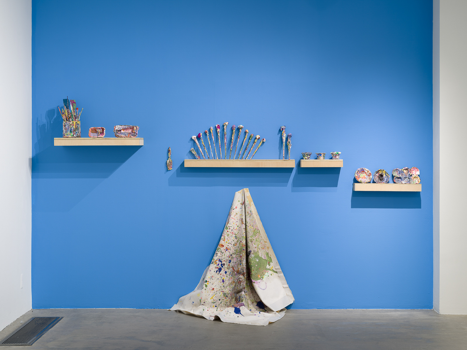 This is a color photograph of a blue wall in an exhibition space of Vanessa McConnell's artworks at the Los Angeles Municipal Art Gallery. There are four floating shelves mounted to the wall, and on each shelf lies paint-covered ephemera, including paint brushes, cups, dishes, and mixing trays. In the center of the wall under one of the shelves is a light colored paint slpattered drop cloth, hung from a single hook draped against the wall and resting on the floor.
