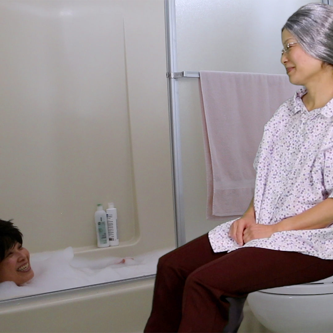 This is a still image from the digital video artwork Bathroom Stall Tears Part 1 by artist Yoshie Sakai. In the image, Sakai is dressed as a younger version of herself (pictured on the left) and her grandmother (pictured on the right). The character on the left is situated in a bathtub with only her head visible and two bottles of soap placed on a small crevice of the wall. She is smiling while looking up at the character on the right. The character on the right is sitting on top of a toilet, and is dressed wearing a short sleeve patterned blouse, dark brown pants, and silver wig styled in a low bun. She has her hands folded in her lap with a slight smile on her face as she looks down at the character in the bathtub. To her right is a white cabinet, and to her left is a light pink colored towel.